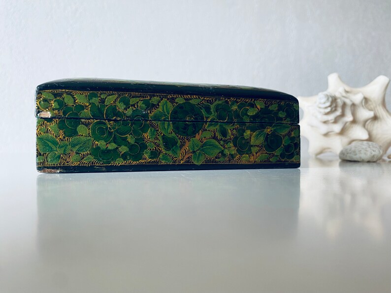 K&ouml;nnte beinhalten: Eine schwarze Holzkiste mit einem gr&uuml;nen und goldenen Blumendesign. Die Kiste ist geschlossen und steht auf einer wei&szlig;en Oberfl&auml;che.