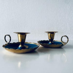 Puede incluir: Dos candeleros vintage de latón con detalles de esmalte azul. Cada candelero presenta una base festoneada, un asa y una copa central para la vela. Los candeleros tienen un acabado dorado y se exhiben sobre una superficie blanca.