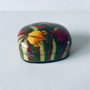 May include: A small, oval-shaped box with a hand-painted design of colorful flowers and leaves on a green background. The box is made of papier-mâché and has a dark brown rim.