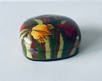 May include: A small, oval-shaped box with a hand-painted design of colorful flowers and leaves on a green background. The box is made of papier-mâché and has a dark brown rim.