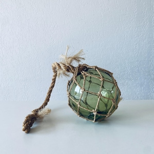 Op de afbeelding: Een groene glazen visserssloep met een bruin touwnet. De sloep is rond en heeft een touwhandvat.