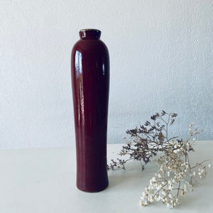 Könnte beinhalten: Eine hohe, dunkelbraune Keramikvase mit abgerundetem Boden und leicht ausgestellter Öffnung. Die Vase steht auf einer weißen Oberfläche.