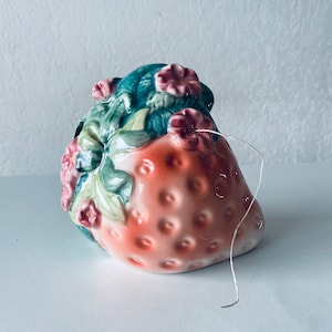 May include: A ceramic strawberry-shaped thread holder with green leaves and pink flowers. The holder has a white thread coming out of it.