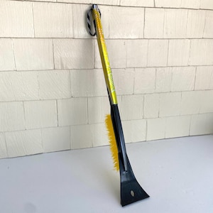 May include: A yellow and black ice scraper with a brush. The scraper has a black plastic blade and a yellow brush. The handle is yellow with black accents and a black wrist strap. The tool is designed for removing ice and snow from vehicles.