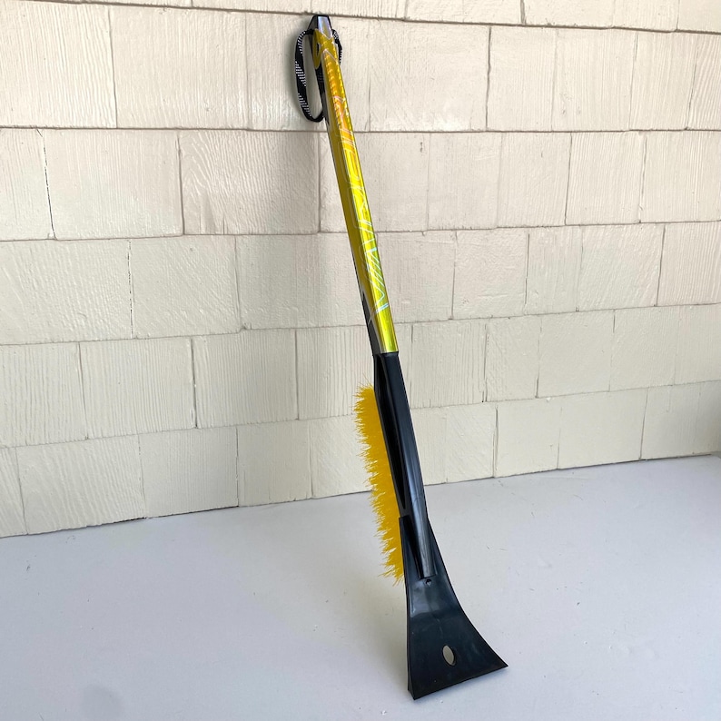 May include: A yellow and black ice scraper with a brush. The scraper has a black plastic blade and a yellow brush. The handle is yellow with black accents and a black wrist strap. The tool is designed for removing ice and snow from vehicles.