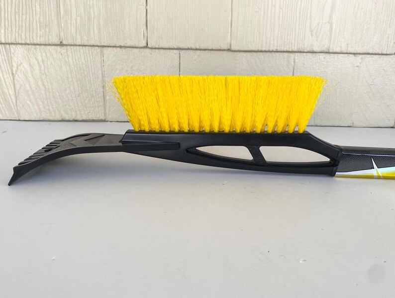 May include: A black and yellow snow brush with a yellow brush head and a black scraper blade. The brush has a long handle for reaching across a car windscreen. The brush is designed for removing snow and ice.