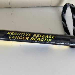 May include: A black hockey stick with the text "REACTIVE RELEASE LANCER R&Eacute;ACTIF" in yellow. The stick has a black and white checkered strap at the end.