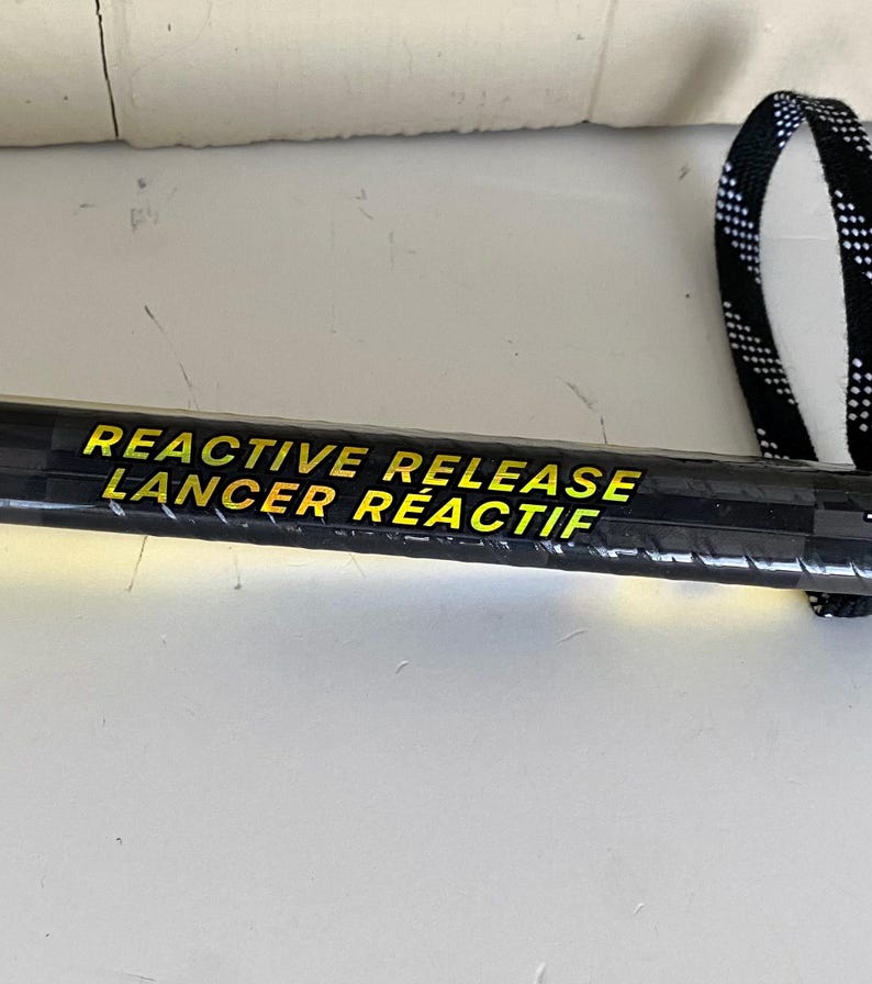 May include: A black hockey stick with the text "REACTIVE RELEASE LANCER R&Eacute;ACTIF" in yellow. The stick has a black and white checkered strap at the end.