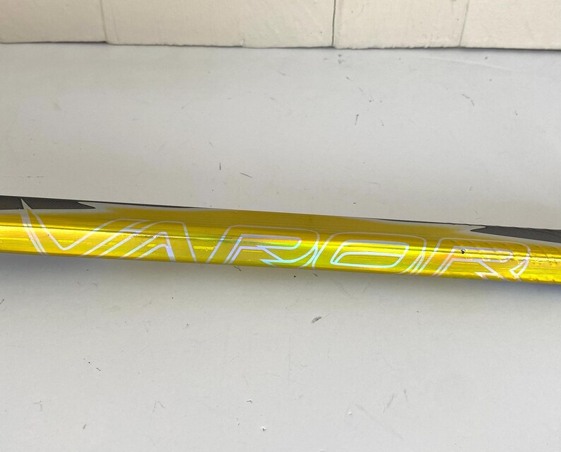 May include: A close-up of a yellow hockey stick with the word "VAPOR" printed in white. The stick has a black accent on the top edge. The background is a plain white surface.