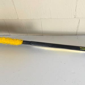 May include: A black and yellow ice scraper with a long handle. The scraper has a black blade with teeth and a yellow brush. The handle has the text "REACTIVE RELEASE LANCER REACTIF". A black and white loop is at the end of the handle.
