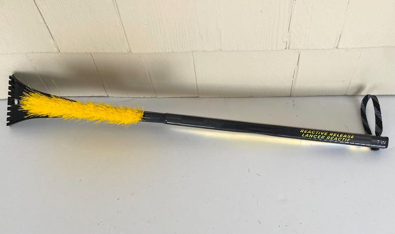 May include: A black and yellow ice scraper with a long handle. The scraper has a black blade with teeth and a yellow brush. The handle has the text "REACTIVE RELEASE LANCER REACTIF". A black and white loop is at the end of the handle.