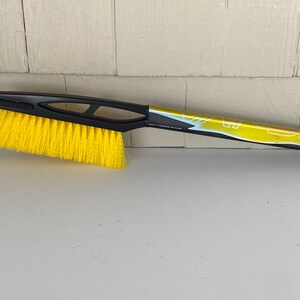 May include: A black and yellow ice scraper and snow brush combination tool. The brush has bright yellow bristles and a black handle with a built-in scraper blade. The handle has a loop for hanging.