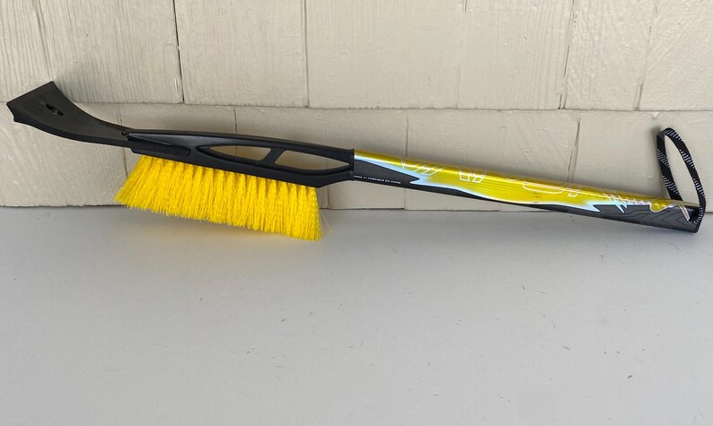 May include: A black and yellow ice scraper and snow brush combination tool. The brush has bright yellow bristles and a black handle with a built-in scraper blade. The handle has a loop for hanging.