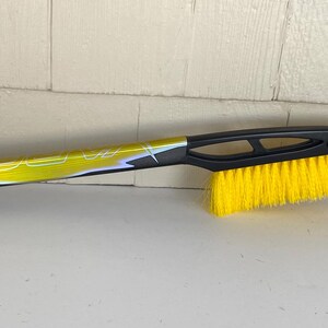May include: A black and yellow snow brush with a yellow brush head and a black ice scraper. The handle is yellow and black with a loop for hanging.