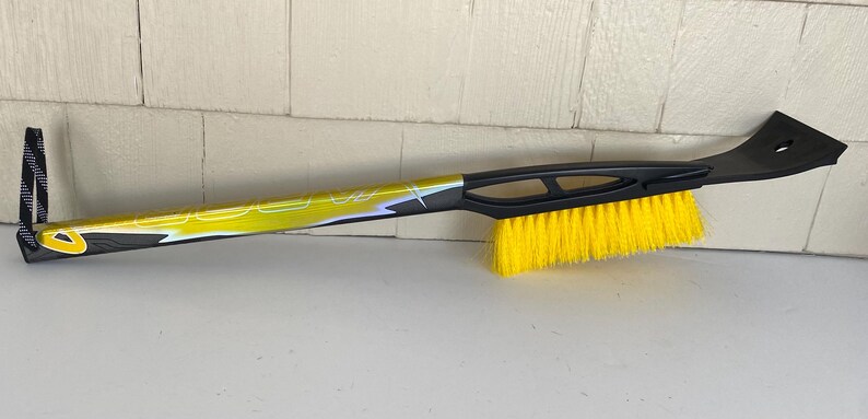 May include: A black and yellow snow brush with a yellow brush head and a black ice scraper. The handle is yellow and black with a loop for hanging.