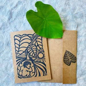 May include: A brown card with a black and white linocut print of a person in a womb-like shape. The print is surrounded by a design of leaves and a sun. The card is part of a set with a matching brown envelope.