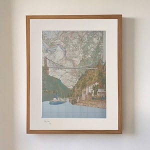 May include: A framed print of a vintage map with a bridge and a boat in the foreground. The map is in shades of brown, green, and blue. The bridge is a suspension bridge with a white cable. The boat is a small, white ferry. The print is signed "Chris Jury 2020".