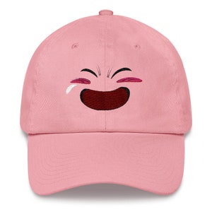 May include: A pink baseball cap with an embroidered cartoon face with a wide smile and tear coming from the eye.