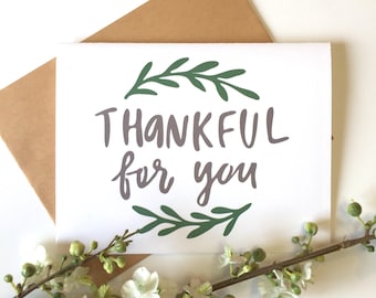 May include: A white card with a green leafy wreath around the words "THANKFUL for you" in gray lettering.