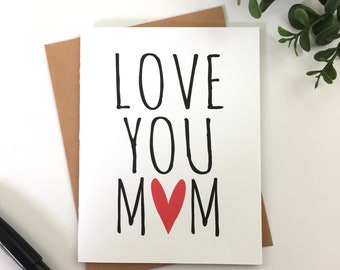 Könnte beinhalten: Weiße Grußkarte mit dem schwarzen Schriftzug "LOVE YOU MOM", wobei ein rotes Herz das "O" in MOM ersetzt. Die Karte befindet sich in einem braunen Umschlag.