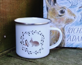 Può includere: Una tazza di smalto bianco con un'illustrazione in bianco e nero di un coniglio che legge un libro all'interno di una ghirlanda di foglie. La tazza è accanto a un libro intitolato "Watership Down" di Richard Adams.