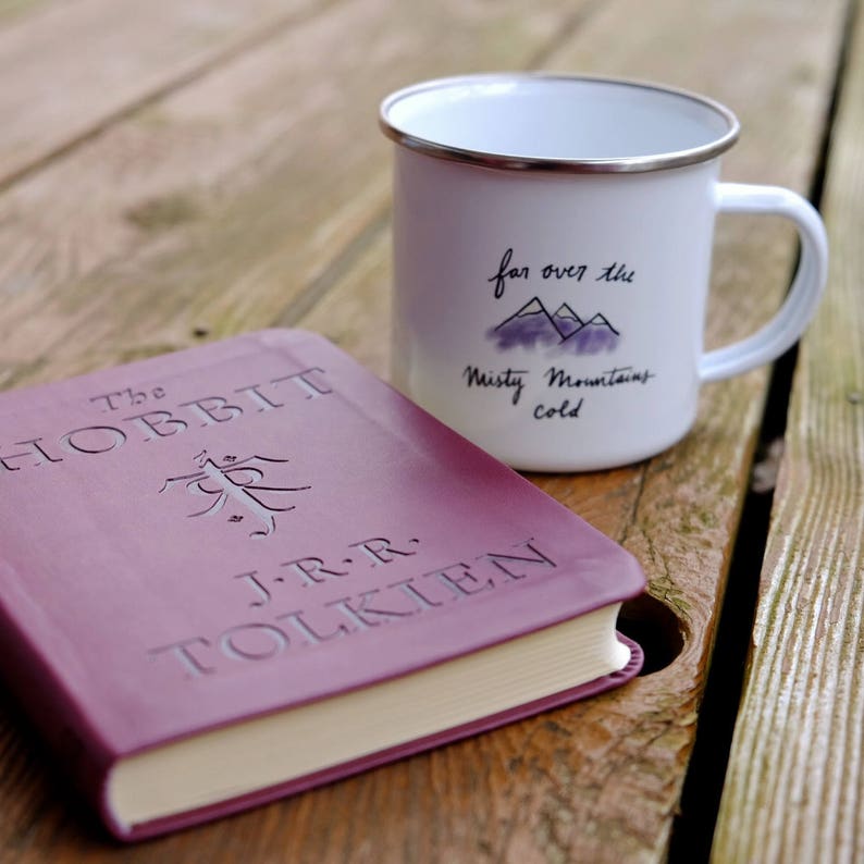 Pu&ograve; includere: Un libro rilegato in bordeaux dal titolo "Lo Hobbit" di J.R.R. Tolkien. Il libro &egrave; appoggiato su un tavolo di legno accanto a una tazza di smalto bianco con il testo "far over the Misty Mountains cold".