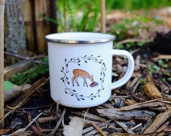 Può includere: Tazza di smalto bianco con un'illustrazione di un cervo marrone all'interno di una ghirlanda di foglie verdi. Il cervo sta guardando un mucchio di libri.
