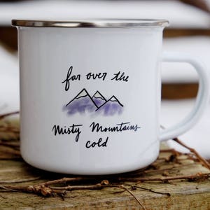 Pu&ograve; includere: Tazza da campeggio in smalto bianco con una citazione scritta a mano in nero che recita "far over the misty mountains cold" e un'illustrazione ad acquerello viola di montagne.