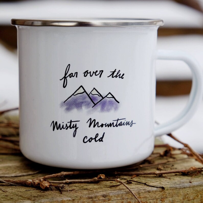 Pu&ograve; includere: Tazza da campeggio in smalto bianco con una citazione scritta a mano in nero che recita "far over the misty mountains cold" e un'illustrazione ad acquerello viola di montagne.