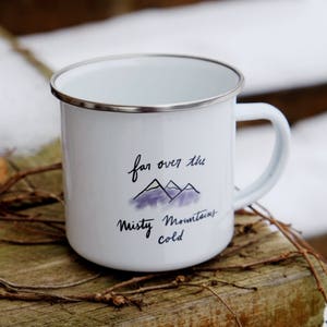 Pu&ograve; includere: Tazza da campeggio in smalto bianco con un'illustrazione di montagna nera e viola e il testo "far over the misty mountains cold".