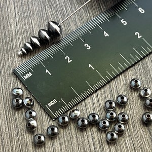 May include: A close-up of a ruler measuring in inches and millimeters, with a group of small, round, silver-colored beads scattered around it.