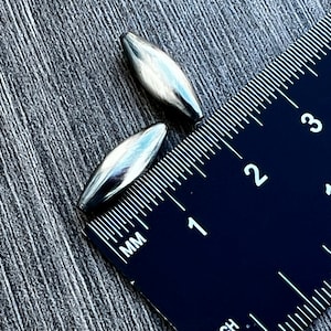 May include: Two silver metal bullet-shaped beads, measuring approximately 2.5 cm in length, are placed next to a ruler. The ruler is marked in millimetres and inches.