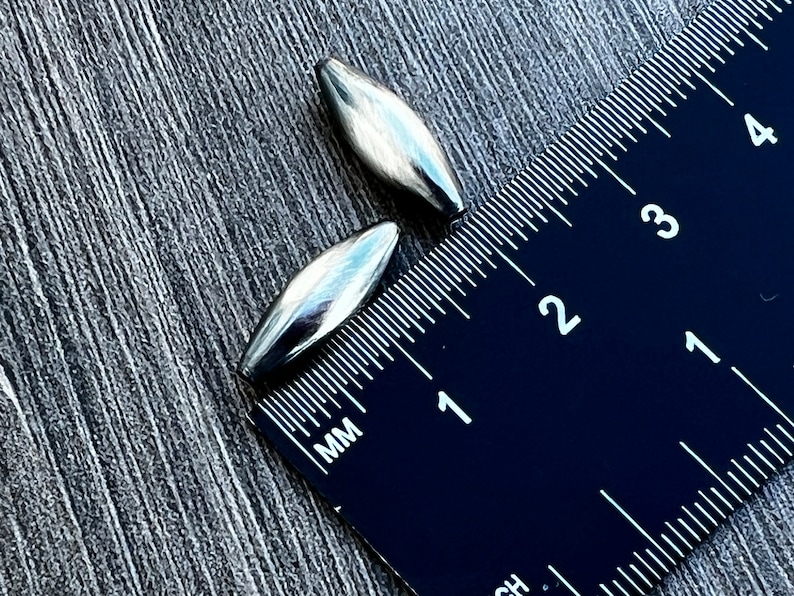 May include: Two silver metal bullet-shaped beads, measuring approximately 2.5 cm in length, are placed next to a ruler. The ruler is marked in millimetres and inches.