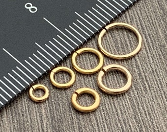 May include: A set of five gold jump rings of varying sizes. The rings are arranged on a black and white surface.