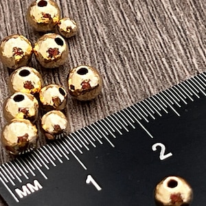May include: A close-up of a group of gold-colored metal beads. The beads are round and have a hammered texture. A ruler is in the background, with the numbers 1 and 2 visible.