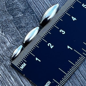 May include: Four silver metal bullet-shaped beads, measured against a black ruler with white markings. The beads range in size from 1.25 cm to 2.5 cm in length.