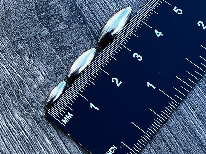 May include: Four silver metal bullet-shaped beads, measured against a black ruler with white markings. The beads range in size from 1.25 cm to 2.5 cm in length.