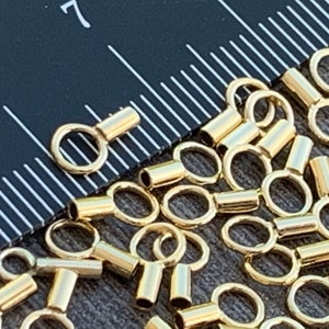 May include: A close-up of a group of gold-colored metal crimp beads. The beads have a small loop on one end and a cylindrical shape on the other end.