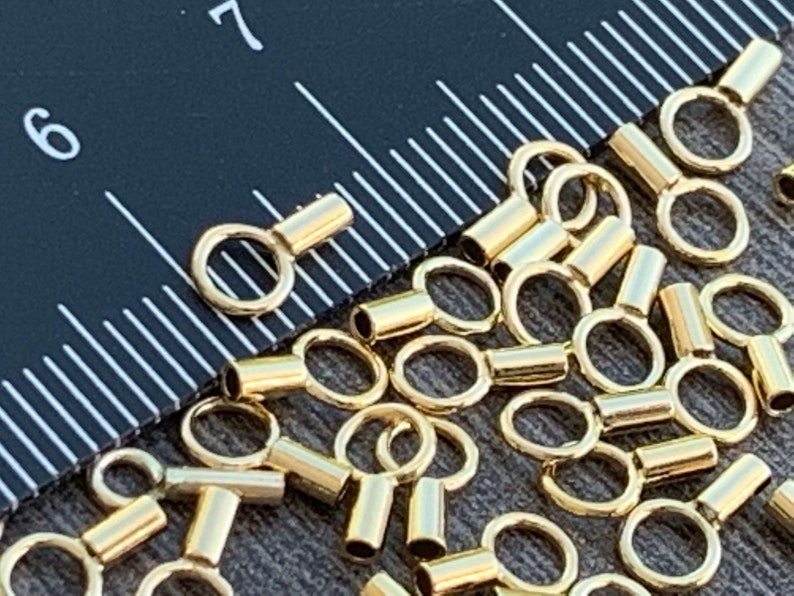 May include: A close-up of a group of gold-colored metal crimp beads. The beads have a small loop on one end and a cylindrical shape on the other end.