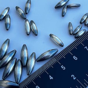 May include: A close-up of a group of silver and grey oval-shaped beads. The beads are arranged on a white surface with a black ruler in the background. The ruler is marked with numbers from 2 to 8.