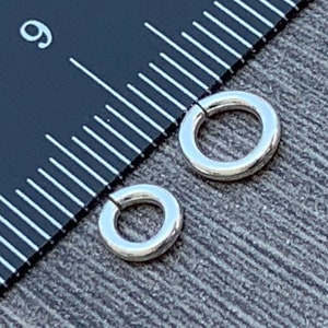 May include: Two silver jump rings, one larger than the other, are shown next to a ruler. The larger jump ring measures 1/2 inch in diameter.