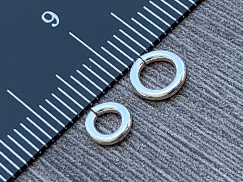 May include: Two silver jump rings, one larger than the other, are shown next to a ruler. The larger jump ring measures 1/2 inch in diameter.