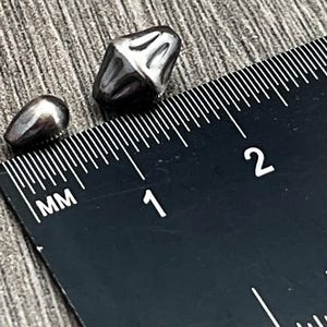 May include: Two silver-colored metal beads, one round and one pointed, are shown next to a ruler marked in millimeters. The round bead measures approximately 1 centimeter in diameter, and the pointed bead measures approximately 1.5 centimeters in length.