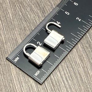 May include: Two small silver-colored metal padlocks placed on a black ruler. One padlock is closed, and the other is open. The ruler shows measurements in inches and millimeters, indicating the padlocks are approximately 0.5 inches in width.