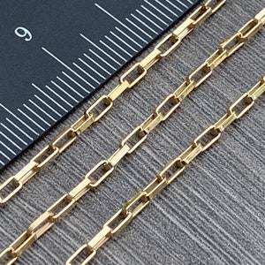 May include: A close-up of a gold chain necklace with a box chain design. The chain is laid out on a ruler with markings in inches.