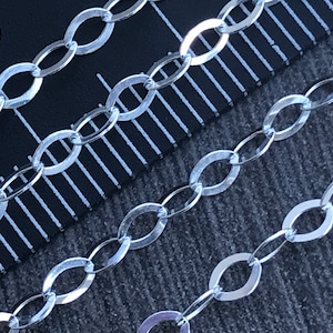 May include: Close-up of a silver chain with oval links. The chain is laid out on a ruler with black and white markings. The chain is approximately 25 cm long.