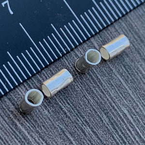 May include: Three silver metal tube-shaped beads, each with a hollow center, are arranged on a gray textured surface. A black ruler with white markings is visible in the background.