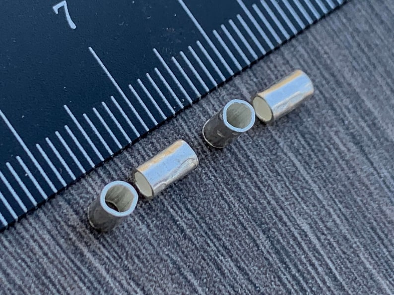May include: Three silver metal tube-shaped beads, each with a hollow center, are arranged on a gray textured surface. A black ruler with white markings is visible in the background.