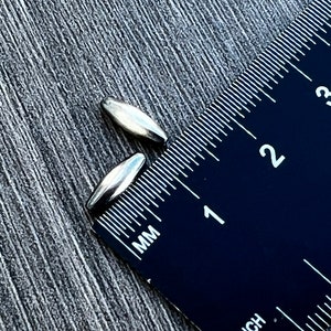May include: Two silver-coloured metal beads, each measuring approximately 2.5 cm in length, are placed on a black ruler with white markings. The ruler is marked in inches and millimetres.