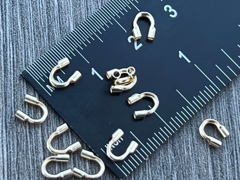 Op de afbeelding: Gouden metalen krimpkralen voor het maken van sieraden. De kralen worden getoond naast een liniaal, waarbij de grootste kraal ongeveer 1,2 cm in diameter is.